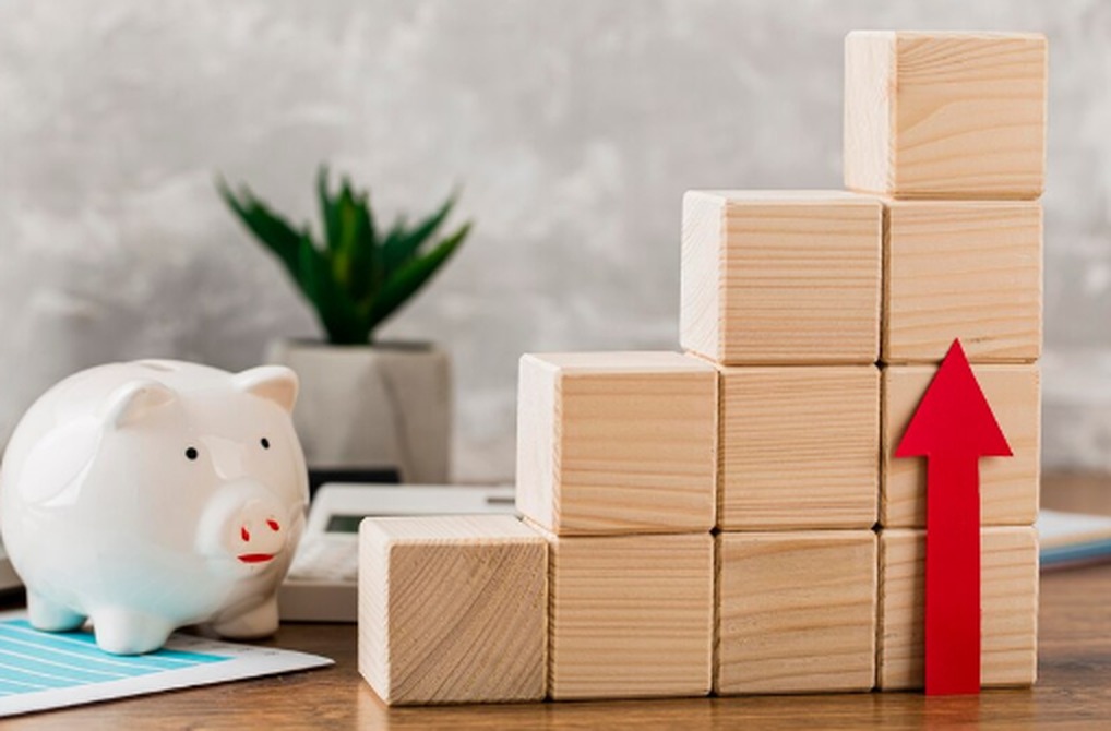 A white piggy bank beside stacked wooden blocks arranged in ascending order, with a red upward arrow pointing to growth, symbolizing how Australian company dividends work to steadily build investor wealth.