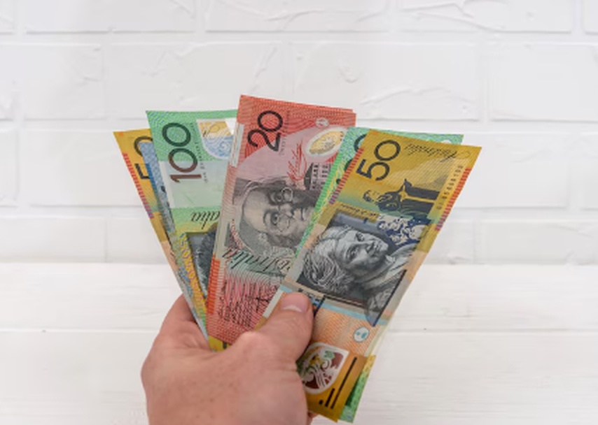 A hand holding a fan of Australian banknotes in front of a white brick background, representing personal investment, cash savings, or participation in the Australian direct treasury system.