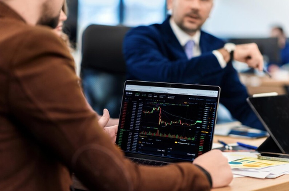 Investor analyzing ETF performance on a laptop screen displaying detailed financial charts and candlestick graphs, while discussing market trends in a professional office setting. The image illustrates real-time decision-making in ETF trading.