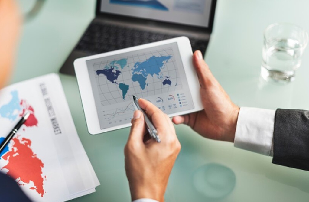 Close-up of a businessperson analyzing a world map with financial data on a tablet, with charts and documents nearby. The scene represents a strategic discussion about investing internationally, highlighting global market trends and regional performance.