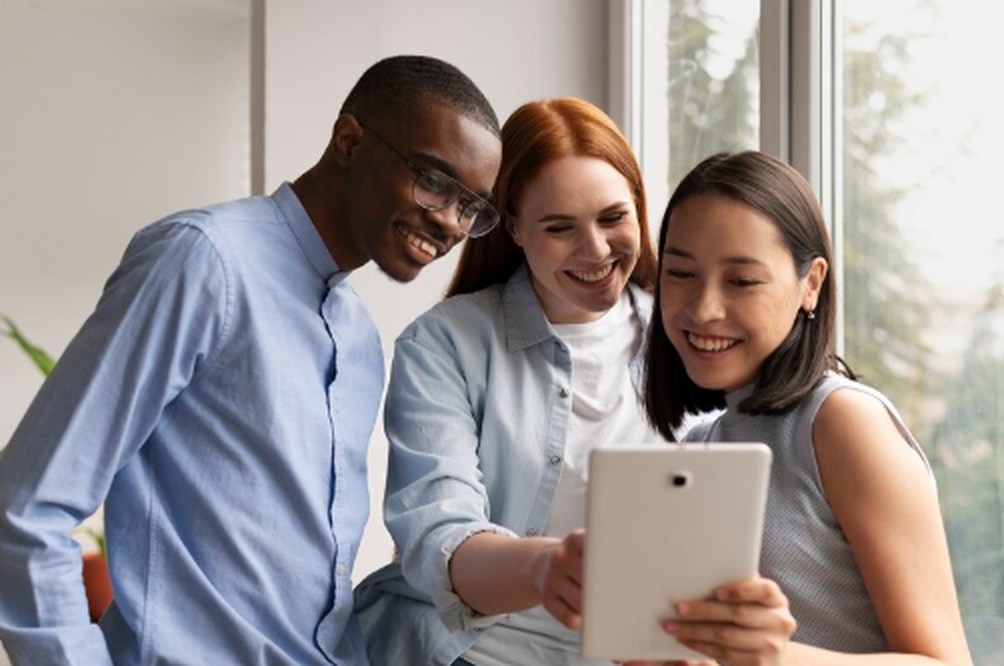 Three young adults smiling and looking at a tablet together near a window, representing collaboration, financial awareness, and exploring investments for young Australian adults.