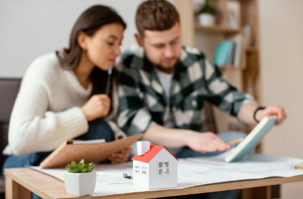 A couple reviews documents at a table with a small model house in the foreground, symbolizing the decision-making process between investing in property or shares.