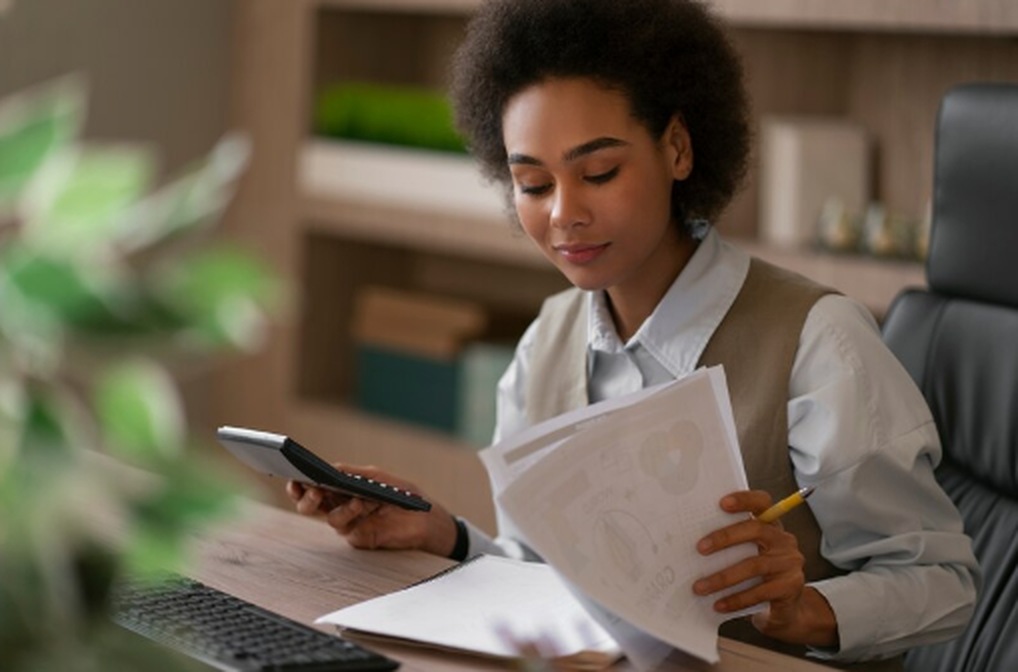 Focused businesswoman sitting at a desk with a calculator and financial documents, reviewing paperwork related to income tax filing. The scene represents careful tax preparation in a professional office setting.