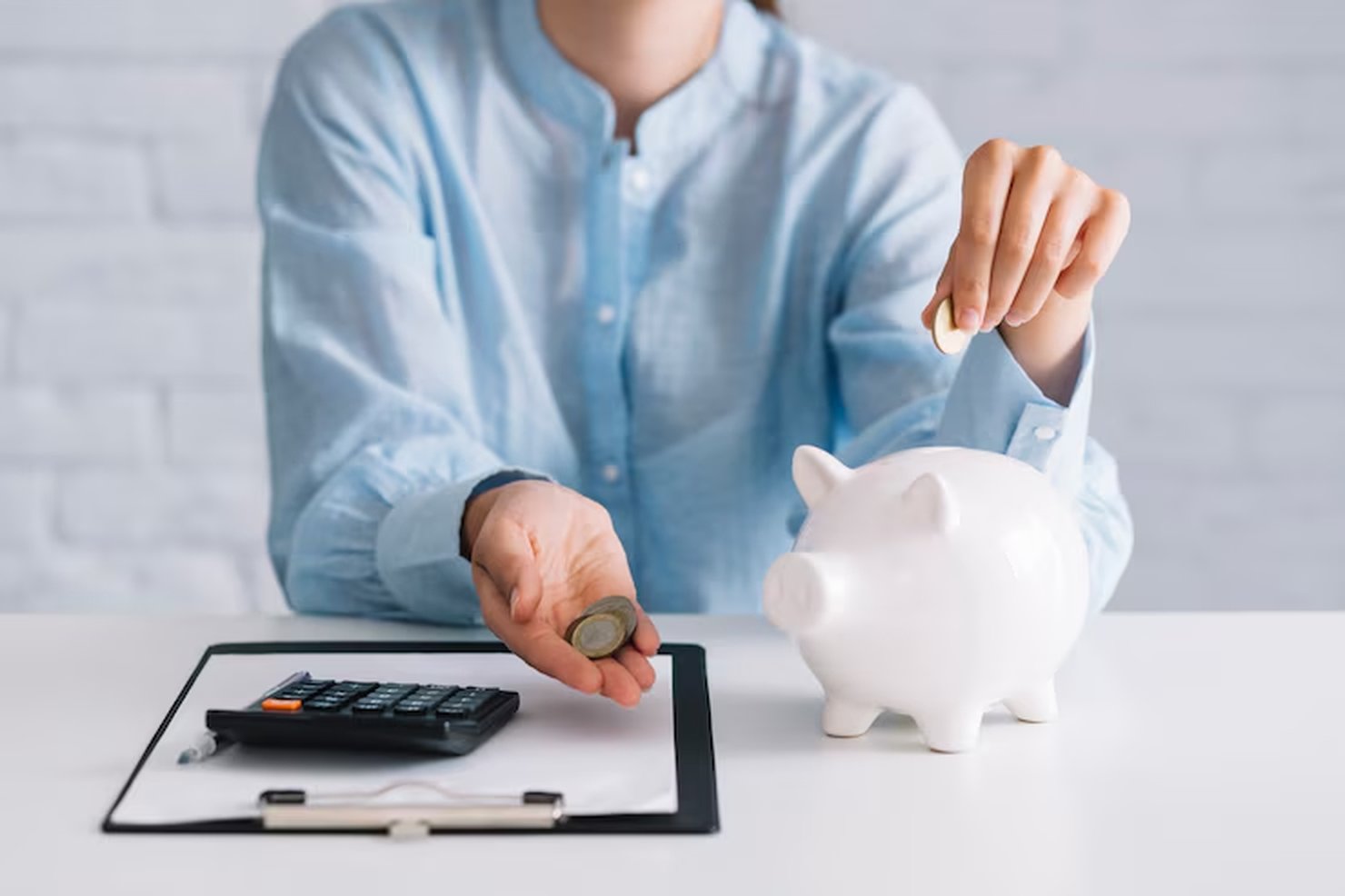 A person in a light blue shirt placing coins into a white piggy bank on a desk, with a clipboard and calculator nearby, illustrating the concept of how to invest with little money through consistent saving habits.