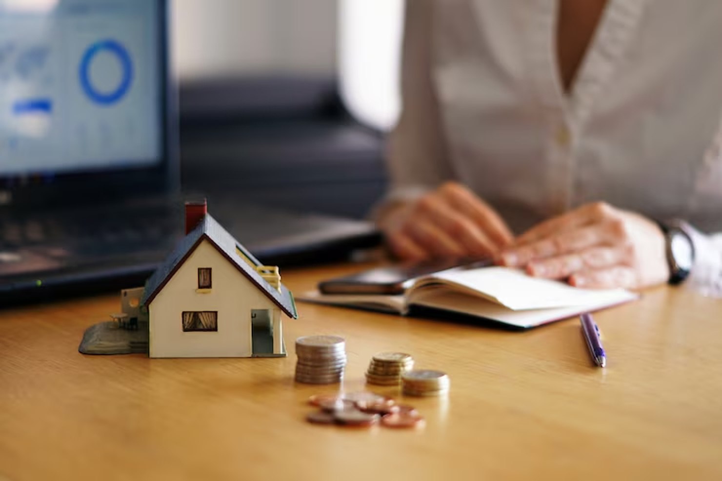 A miniature house model placed next to stacked coins on a wooden desk, with a person writing in a notebook and a laptop displaying financial data in the background, symbolizing the concept of investing in real estate.