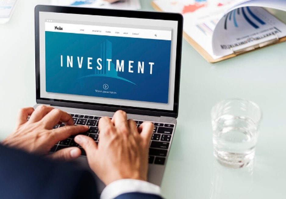 Close-up of a person typing on a laptop with the word “INVESTMENT” displayed on the screen, suggesting online research or management of investment funds. A glass of water and printed documents are visible on the desk, reinforcing a professional setting.