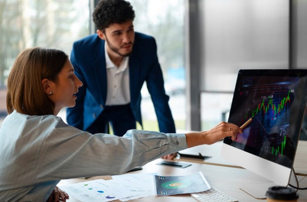 Businesswoman pointing at a financial chart on a desktop monitor while discussing market trends with a male colleague in a modern office. They appear to be analyzing data related to mistakes in investments, with printed reports and graphs visible on the desk.