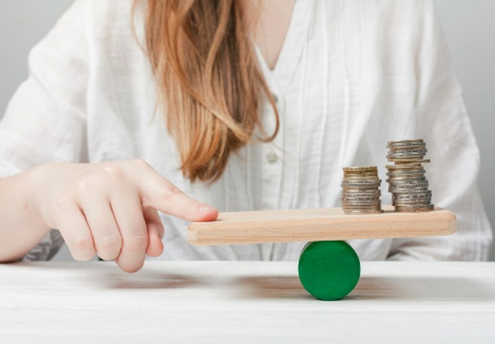 A person balances stacked coins on a wooden board supported by a green cylinder, symbolizing the concept of financial stability and strategies to protect your portfolio against inflation.