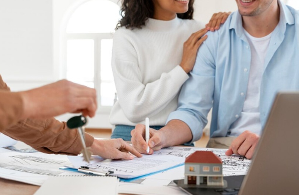 A smiling couple signing documents at a desk with a miniature house model, keys, and architectural plans, representing the concept of investing in real estate funds and property ownership.
