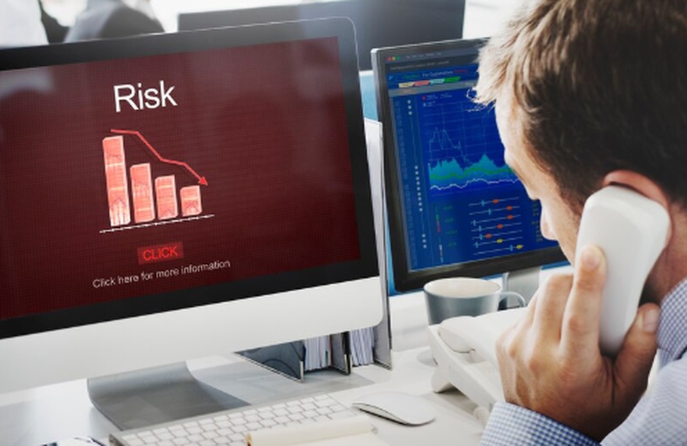 A man on the phone looks at computer monitors displaying financial charts and a warning screen labeled "Risk," illustrating the analysis of the risk of an asset in investment decision-making.