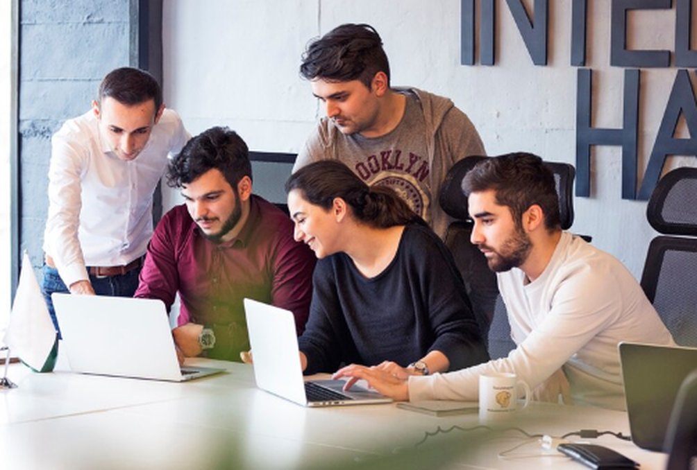 A diverse group of young professionals collaborating around laptops in a modern office setting, symbolizing teamwork and innovation within the startup ecosystem. The image captures a dynamic moment of brainstorming and tech-driven problem solving often seen in startups.