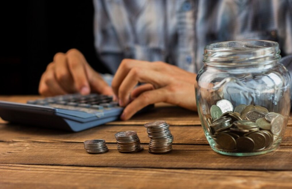 A person calculating coins with a jar full of loose change on a wooden table, representing careful personal savings and financial planning related to superannuation funds.